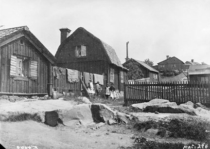 Svart-vit bild fotograferad uppåt en bergsknall. I mitten syns barn framför ett trähus och ett plank där det hänger tvätt. I förgrunden stenar och jord.