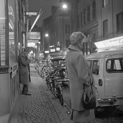 Svartvitt fotografi på kvinna i kappa och handväska på en upplyst trottoar 