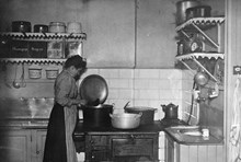 Interiör från köket på Katarina västra barnkrubba med en kokerska som står vid vedspisen och lagar mat.