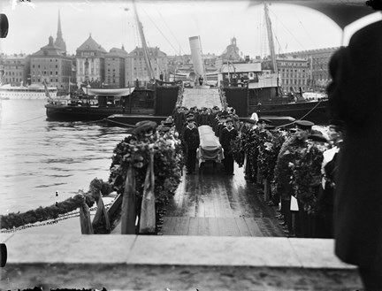 Från en båt förankrad i Stockholms ström, förs en kista iland via en tillfällig spång. Spången kantas av sjömän med blomsterkransar. I bakgrunden syns den äldre bebyggelsen i Gamla stan.