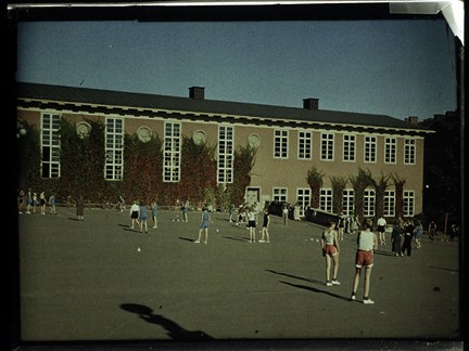 På en skolgård syns många barn i mellanstadieåldern som är klädda i gymnastikkläder, linnen och shorts. I bakgrunden syns en skolbyggnad i tjugotalsklassicistisk stil. 