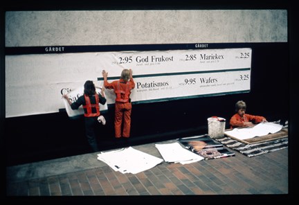 Tre män i orange kläder står på spåren och klistrar upp reklamaffischer.