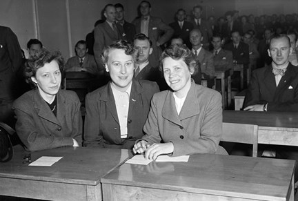 Tre kvinnor sitter bakom två skolbänkar. I bakgrunden sitter de andra eleverna som alla är män.