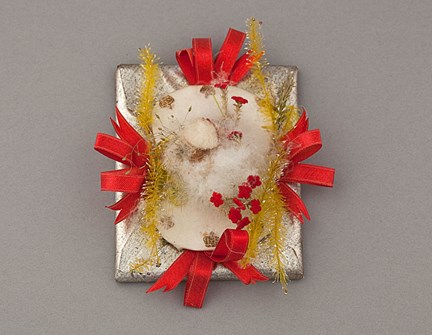 En platt karamell är inslagen i silverfärgat papper. Ovanpå har karamellen dekorerats med röda band, gula glittrande kvistar och små röda blommor.