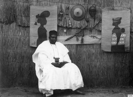 En svart man i vita kläder sitter utanför en vasshydda. På väggen bakom honom hänger bilder och hantverksföremål.