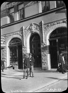 Två män står vid Odengatan 67. Till vänster en bossättningsaffär, Flodins, till höger en cigarrhandel