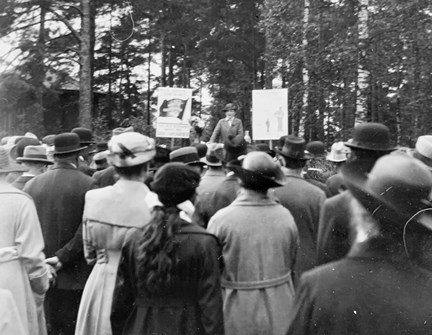 Agda Östlund står i en talarstol i skogsmiljö i Enskede musikpark söndagen den 11 september 1921. Hon har stor publik, där de flesta är kvinnor.