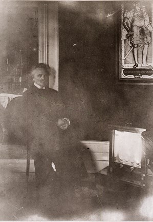 Svartvitt foto där Strindberg sitter på en stol i sin salong och värmer sig vid ett elektriskt element
