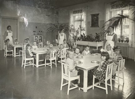 Svartvit bild där mindre barn sitter vid matbord, de har hakklappar och en del har händerna knäppta.