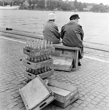 Vy från Skeppsbrokajen mot Skeppsholmen. Två äldre kvinnor sitter och vilar på en tomlåda. Träbackar med tömda Coca Colaflaskor i förgrunden