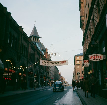 Människor passerar, butiksskyltar syns (kaffe, cigaretter) och Östermalms saluhall skymtar på vänster sida.