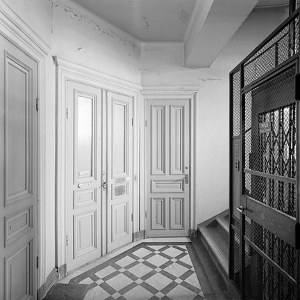 Ett våningsplan i ett trapphus. Golvet är av mönsterlagd marmor. Till höger syns en hiss och dess gallerdörr. Till vänster ett flertal lägenhetsdörrar, både pardörrar och enkeldörrar. 