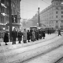 Stureplan och Birger Jarlsgatan söderut. Stureplan 2 och 3. Folk väntar på buss/spårvagn i snöyran. På två stolpar sitter affischer med ett snapsglas och ett ljus, ""Släck inte julglädjen""