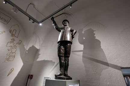 En skulptur i koppar. Den högra armen är höjd med ett ris i handen. Statyn står i ett trapphus