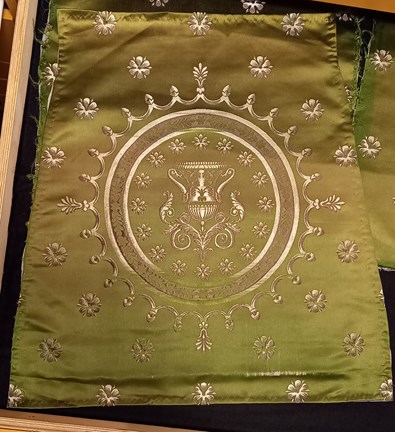 Ett grönt tyg med detaljer i vitt. Runt om finns enkla vita blommor. I tygets mitt finns en rund ram som innesluter två mindre cirklar. Längst in likadana blommor som i resten av tyget runt en urna med handtag. 