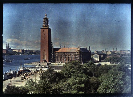 Färgbild med Stockholms stadshus i centrum. Byggställningar syns fortfarande runt huset. I nedre delen av bilden syns grönskande träd och till vänster Mälarens vatten.