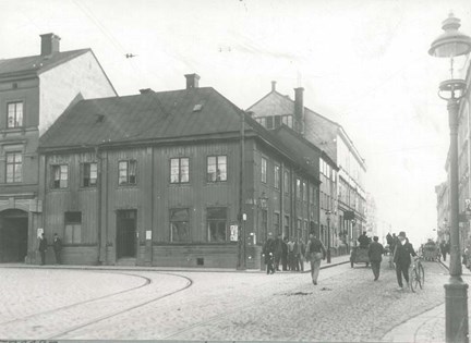 Folkliv framför trähuset där krogen låg. Här skymtar en polis, en man med cykel, ett par kärror, med mera.