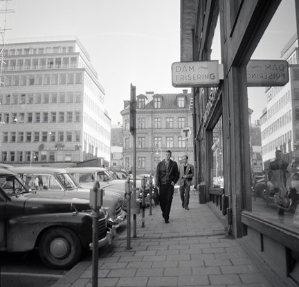 Män går på trottoaren med skyltfönster till höger och parkerade bilar till vänster.