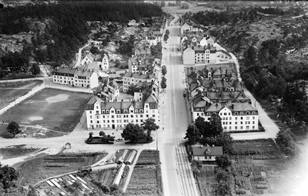 Flygfoto över Aspudden, stenhus i jugendstil längs med stor bilväg, grönområde och skogslandskap runt om