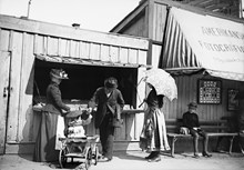 Marknadsstånd på Skansen med marknadsbesökare och barn i skrinda. 
Till höger under markisen har "Amerikanska - Fotografi Ateliern" sin fotoateljé.