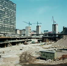 Utgrävd grop med barack, byggmaterial och bråte på platsen för nuv. Sergels Torg. Vy mot femte Hötorgshuset och bankbygge i kv. Hästskon