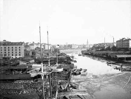 En kanal med bebyggelse på båda sidor. Längst fram master från en båt.