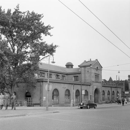 En tvåvåningsbyggnad med valvbågar i gatuplanet. En bil står parkerad på gatan framför huset. I bildkant ett stort träd med tre män gående under och i luften luftledningar.