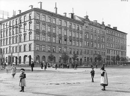 Ett kvarter med tre femvåningshus sett från andra sidan gatan. Barn och vuxna rör sig framför husen.