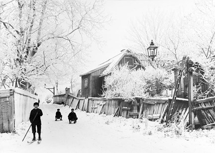 Vinterbild. ett fallfärdigt plank till höger med ett gammalt trähus bakom. I backen åker barn kälke och en pojke åker skodor.