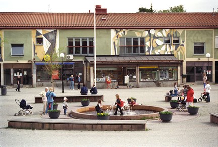 Vuxna och barn rör sig på torget. I mitten en fontän.