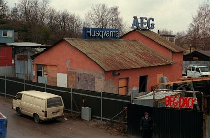En parkerad folkvagnsbuss framför ett högt staket med slitna byggnader bakom.