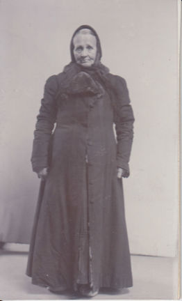 Kvinna klädd i sjalett och lång kappa står och tittar rakt in i kameran