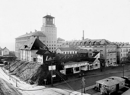 På en liten bergknalle intill en större gatukorsning, ligger några små och ålderdomliga hus kvar. Bakom dem reser sig en stor och nybyggd varuhusbyggnad med torn. Intill ligger en kontorsbyggnad och några bostadshus. I bildens högra hörn ligger en låg kioskbyggnad.