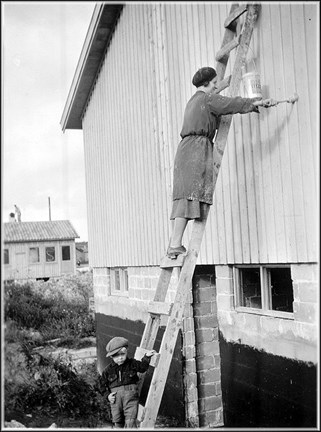 En kvinna står på en stege och målar en husfasad. På marken står hennes son med en hand på stegen och tittar på fotografen. 