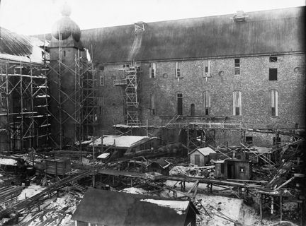 Svart-vit bild från Stadshusets innergård. Murad fasad med tak i bakgrunden. I främre delen av bilden syns små träbaracker, byggställningar och spår för transportvagnar. Snö på marken.