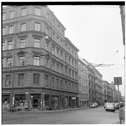 Gatubild med hörnhus med tobaksaffär. På gatan står bilar parkerade. 
