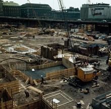 Grund- och byggnadsarbeten i de utgrävda kvarteren Skansen och Frigga. Vy åt söder mot Vattugatan och Drottninggatan på stolpar