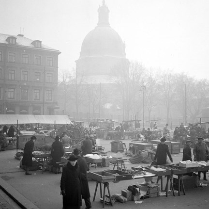 Dimman ligger tät över ett torg. På torget står det många försäljare, en del med tak, andra bara vid bord. I bakgrunden anas en stor kyrkobyggnad och ett äldre flerfamiljshus av sten.