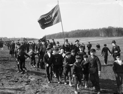 Svart-vitt fotografi med människor i olika åldrar som går i demonstration. Barn syns i förgrunden  och en äldre man i skägg bär en fana med texten "Allmän rösträtt." I horisonten syns skog.