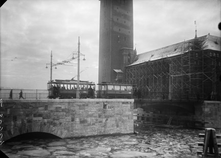 En spårvagn i mitten av bild rör sig framåt över en bra. I förgrunden syns vatten med isflak och i bakgrunden Stadshuset med byggställningar runtom.