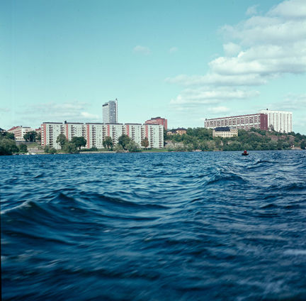 Från en båt på vatten ser vi vågor och i bakgrunden land med flera moderna byggnader. Två låga och breda hus, ett jättehögt och smalt, ett högt och lite bredare. Ett guld, äldre hus syns också till höger. Längs strandkanten växer träd.