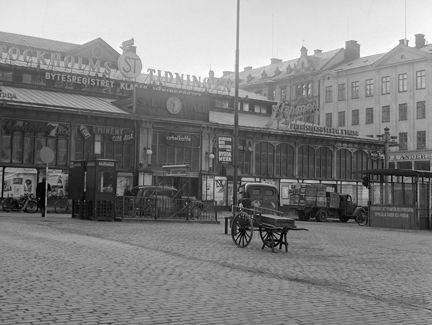 Vid ett torg som är belagt med gatsten ligger en låg saluhallsbyggnad. Saluhallen är av gjutjärn med stora fönsterpartier och en mängd olika reklamskyltar på taket och fasaden. På torget står en telefonkiosk, flera bilar och en kärra. I bakgrunden syns ett par stadshus av sten.