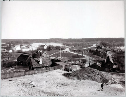 Svart-vit bild med utsikt från en höjd mot byggnader och vatten. I förgrunden till höger syns en liten människofigur.