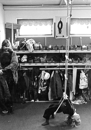 Ett barn gungar i ett kapprum och en kvinna tittar på, 1970-tal