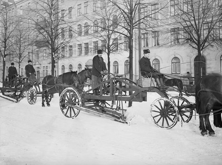 Flera ekipage med hästförspända snöplogar. Kala träd och bebyggelse i bakgrunden.