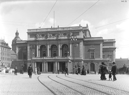 Kungliga operan med spårvagnsspår i förgrunden. Människor promenerar förbi. En kärra i bakgrunden.