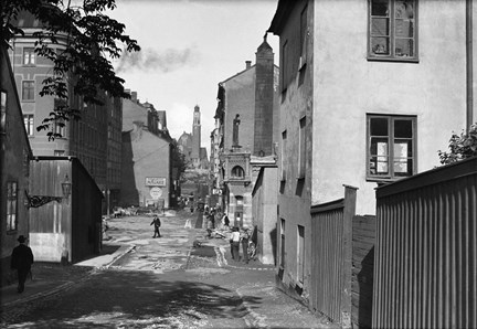 En stadsgata kantad av både större stenhus och äldre små hus. Till höger ett plank. En bit fram håller ett äldre stenhus på att rivas. I slutet av den långa gatan syns en kyrka med ett högt och smalt torn. 