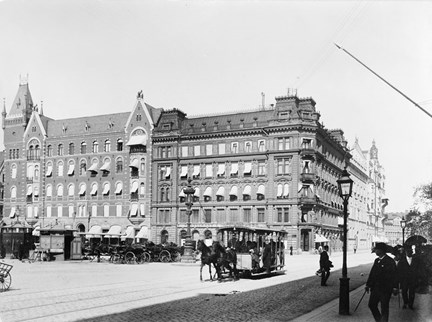 en stor öppen plats, ett rotg, med hästdragen spårvagn. människor flanerar på trottoaren. i bakgrunden stora stenhus med markiser för fönstren.