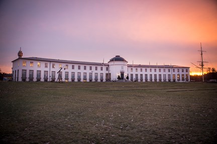 En vit museibyggnad med gräsmatta framför. Bakom museet går solen upp.