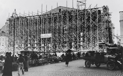 Hus under uppbyggnad, byggnadsställningen i trä framför. I förgrunden Hötorget med några människor och en bild.